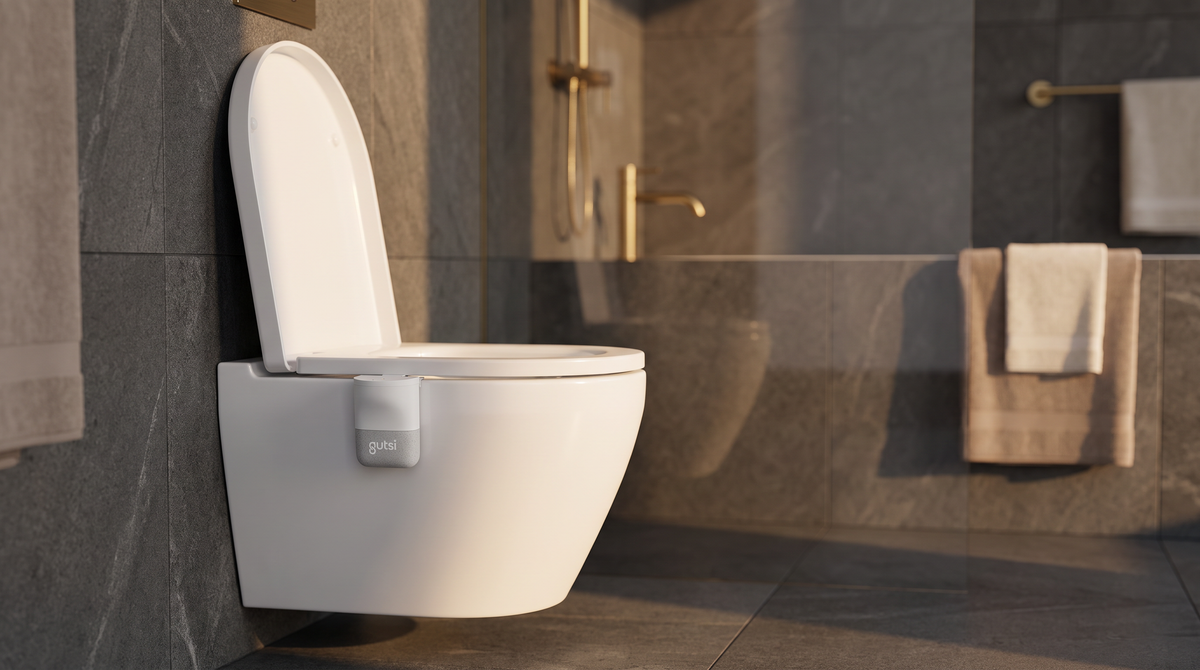 A Gutsi tracker clipped to the rim of a wall-hung toilet in a dark stone bathroom with brass fittings